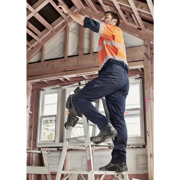 A man in orange and grey high-visibility clothing and navy cargo pants stands on a ladder, working on a ceiling.