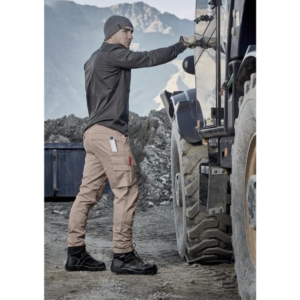 Men's curved cargo pant in beige, featuring multiple pockets, paired with a dark shirt and sturdy boots. Suitable for rugged environments.