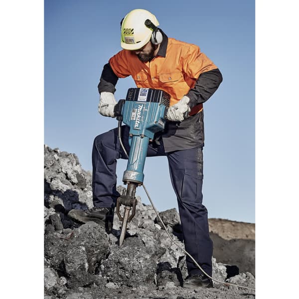 A man in an orange and black work shirt and dark cargo pants operates a jackhammer on rocky terrain.