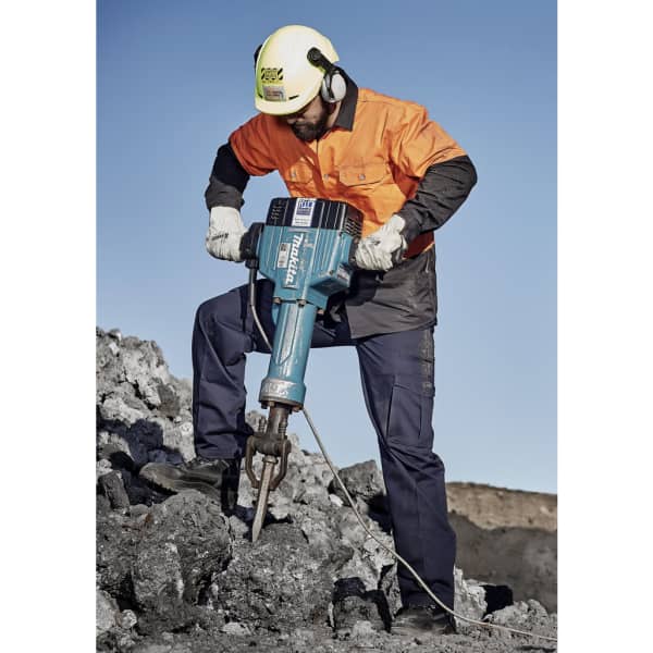 A man in safety gear operates a jackhammer on a rocky surface, wearing an orange shirt and navy blue cargo pants.