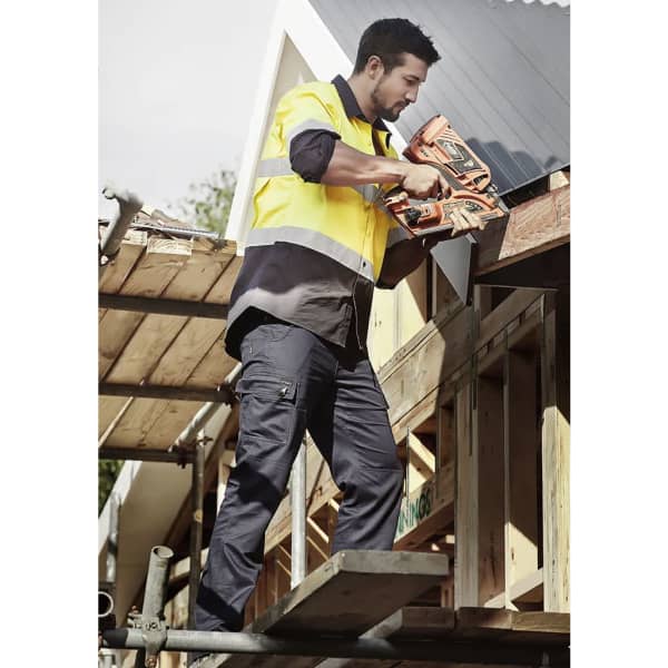 A man wearing a high-visibility shirt and navy cargo pants uses a nail gun on a construction site. The pants have a logo.