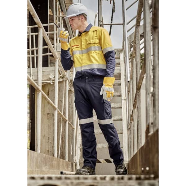 A worker in a yellow and navy work shirt and cargo pants, wearing gloves and a hard hat, on a metal staircase.