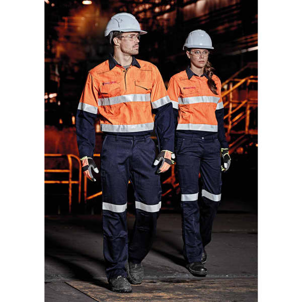 Two workers in high-visibility orange shirts and navy cargo pants, wearing helmets and safety glasses, in an industrial setting.