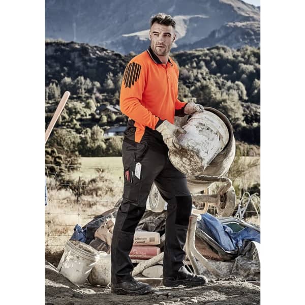A man wearing orange and black work pants stands on a construction site, holding a bucket with tools visible in his pockets.