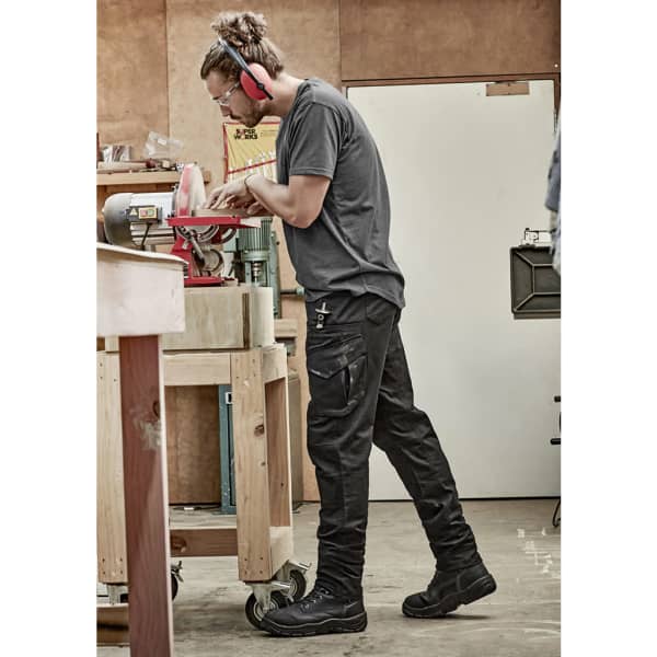 A person wearing black rugged stretch cargo pants, using a tool at a workshop, with hearing protection on.
