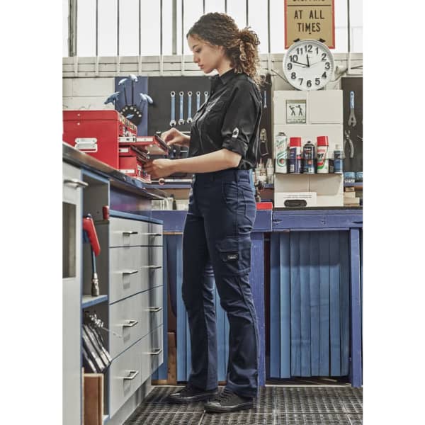 A woman in a black shirt wears navy rugged cargo pants while working in a workshop, surrounded by tools and equipment.