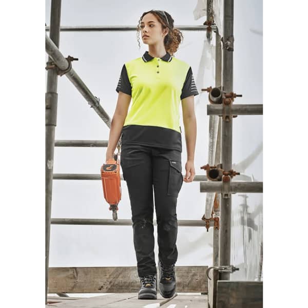 A woman in a high-visibility yellow and black polo shirt and black cargo pants stands on scaffolding, holding a tool.