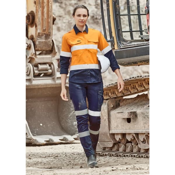 A woman is wearing orange and navy workwear cargo pants with reflective stripes, standing near construction equipment.