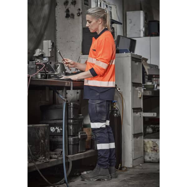 A woman wearing orange and navy cargo pants with cuffs, working at a table in a workshop. The pants feature reflective stripes and a logo.
