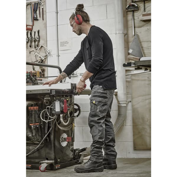 A man wearing black work pants, headphones, and safety glasses operates a machine in a workshop.