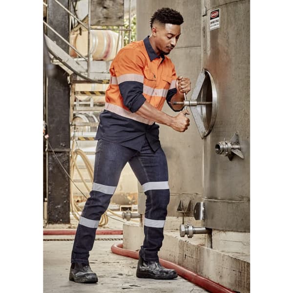 A man in an orange and navy work shirt and blue stretch jeans adjusts equipment, featuring reflective stripes and safety boots.