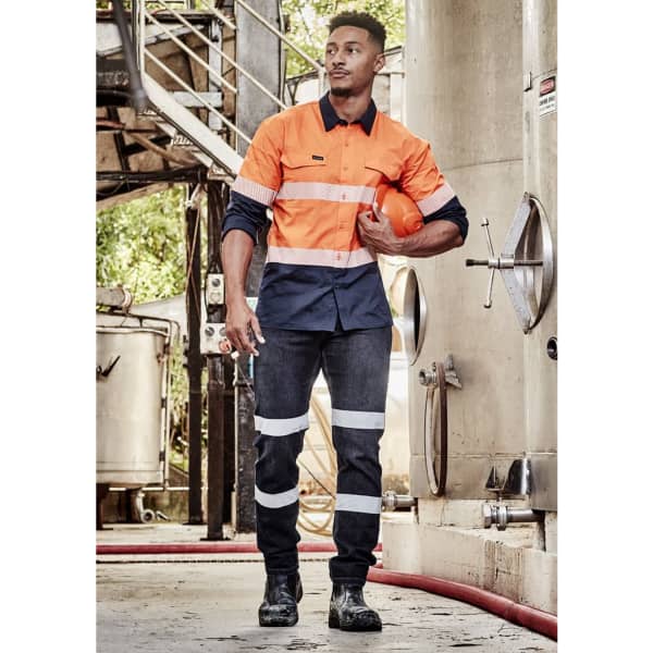 A man in orange and navy workwear with reflective stripes, holding a hard hat, stands in an industrial setting.