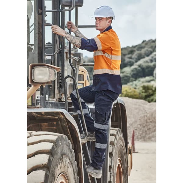 A worker in an orange and navy blue shirt and rugged black pants climbs onto machinery, wearing a safety helmet.
