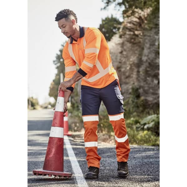 A man wearing high visibility orange and navy work pants adjusts a traffic cone on a rural road.