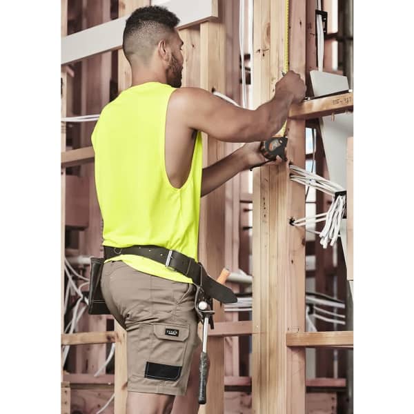 A man wearing a fluorescent yellow sleeveless shirt and beige cargo shorts, working on wooden construction framing.