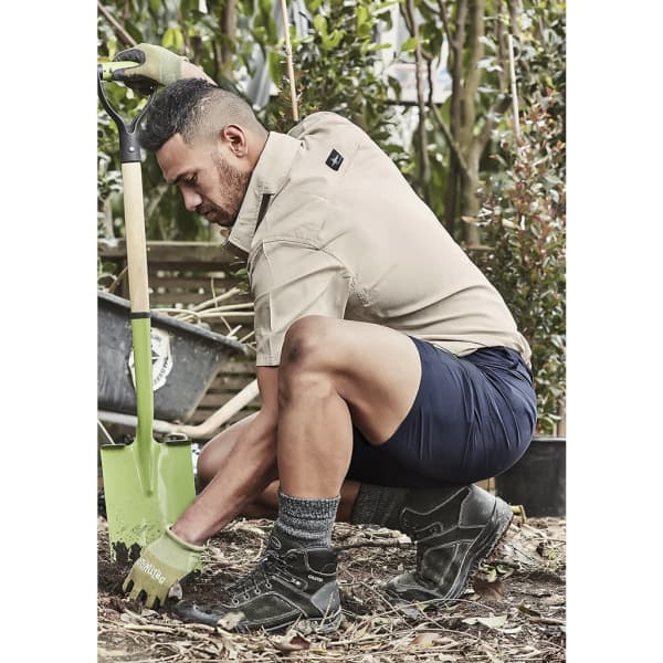 A man kneels in a garden, wearing navy lightweight outdoor shorts and a beige shirt, with a shovel nearby.