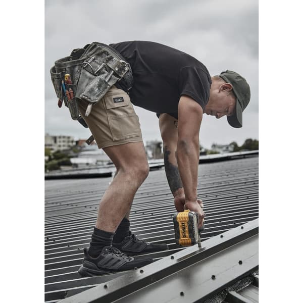 A person wearing khaki cargo shorts and a black t-shirt is using a power drill on a roof. Tools hang from a tool belt.