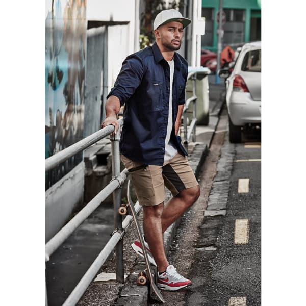 A man wearing a navy shirt and tan stretch shorts, leaning against a railing with a skateboard nearby.