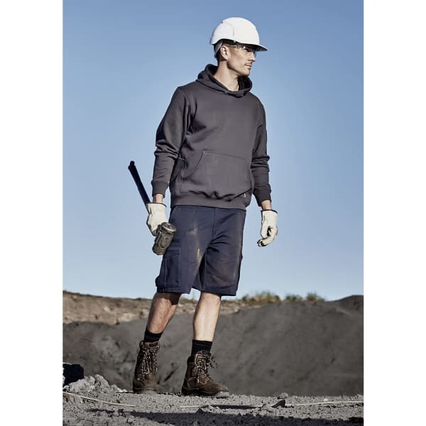 A man wearing a grey hoodie and navy cargo shorts, holding a tool, with a hard hat and work boots in a rugged outdoor setting.