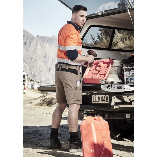 A man in an orange high-visibility shirt and beige shorts stands by a vehicle, holding a drill and tools.
