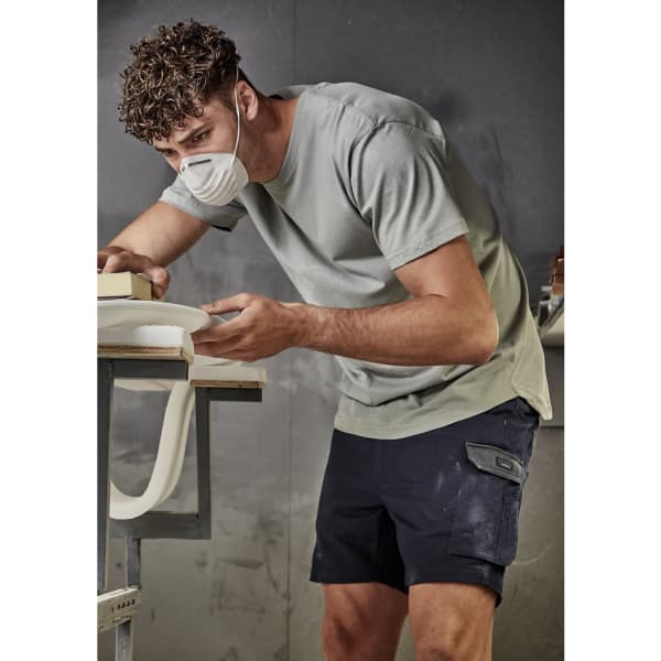 A man wearing a grey t-shirt and navy shorts, working on a project with a mask on in an indoor workspace.
