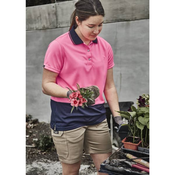 A woman in a pink and navy polo shirt holds flowers, wearing khaki cargo shorts, surrounded by gardening supplies.