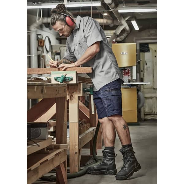 A man is wearing navy work shorts and a light grey shirt while measuring wood in a workshop, equipped with safety gear.