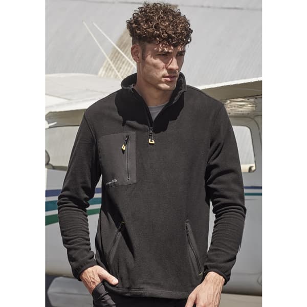 A man wearing a black unisex polar fleece with a quarter-zip and front pockets, set against an aircraft backdrop.