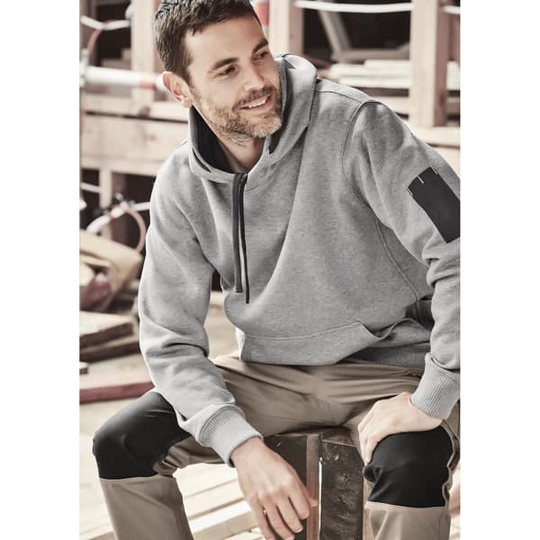 A grey unisex multi-pocket hoodie with a hood, featuring a logo, worn by a man sitting on a wooden structure.