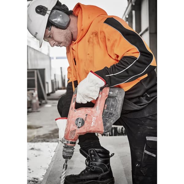 A worker in an orange and black hi-vis pullover hoodie using a power drill while wearing protective gear and gloves.