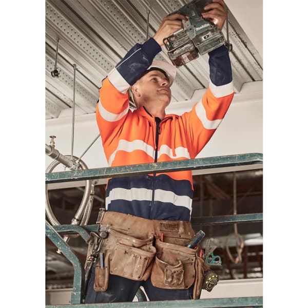 A worker in an orange and navy hi-vis taped hoodie, holding a tool while on a ladder, wearing a hard hat and tool belt.