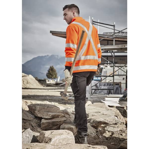 A person wearing an orange hi-vis jumper with reflective stripes, holding a hammer on a construction site.