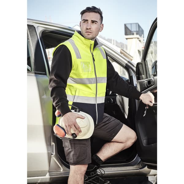 A unisex hi-vis softshell vest in bright yellow and black, with reflective strips, worn by a man stepping out of a vehicle.