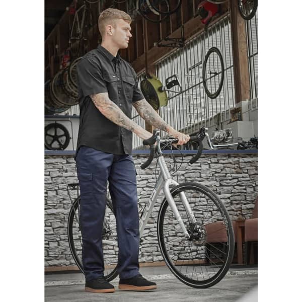 A man in a black shirt and navy cargo pants stands with a bicycle in an indoor setting. The image features industrial decor.