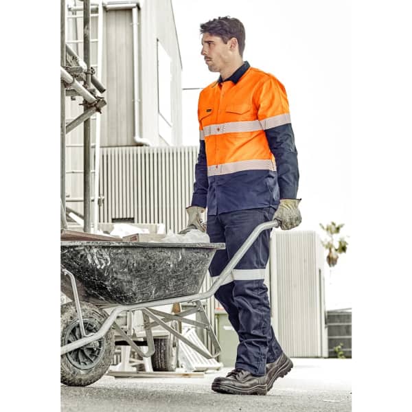 A man in an orange and navy utility pant pushes a wheelbarrow at a construction site. The pants have reflective detailing and a logo.
