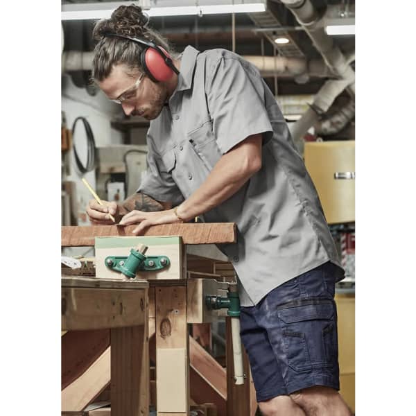 A man in a grey short sleeve work shirt and navy shorts is measuring wood at a workshop, wearing safety glasses and ear protection.