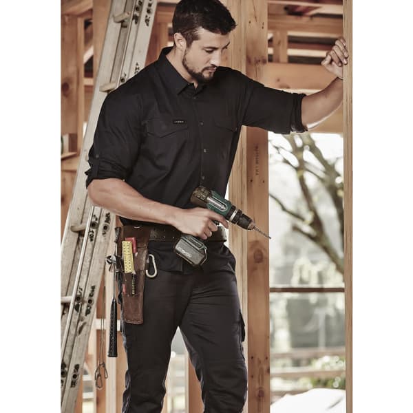 A man in a black long sleeve shirt holds a drill while standing in a wooden frame structure, with tools on his belt.