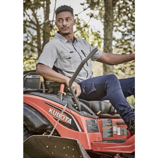 A man wearing a grey short sleeve button-up shirt sits on a red Kubota lawn mower amidst greenery.