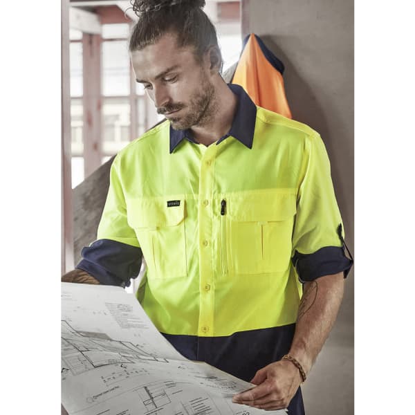 A man in a high-visibility yellow and navy long sleeve work shirt examines a blueprint in an indoor setting.