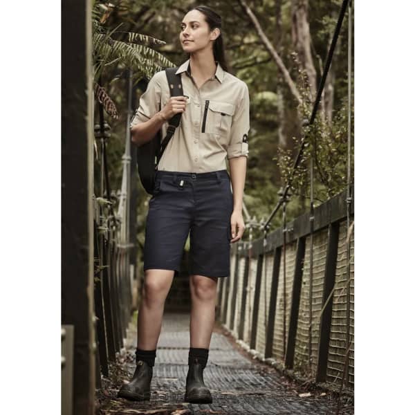A woman in a beige long sleeve button-up shirt and navy shorts stands on a forest walkway, carrying a bag.