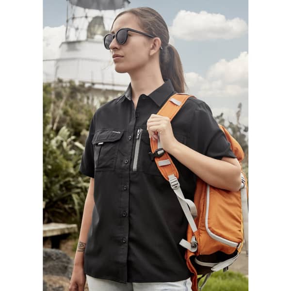 A woman in a black short sleeve outdoor shirt carries an orange backpack, standing near a lighthouse.