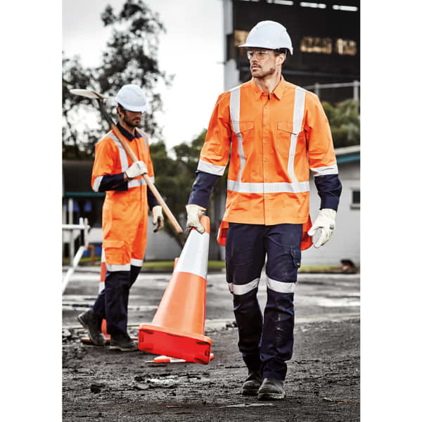A man in an orange rugged work shirt and navy pants carries a safety cone, with another worker in the background.