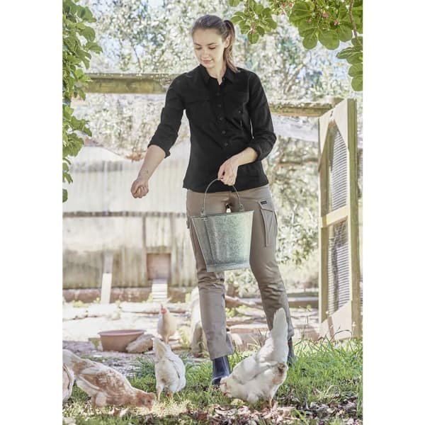 A woman in a black long sleeve shirt and grey pants feeds chickens, holding a bucket in a farm setting.
