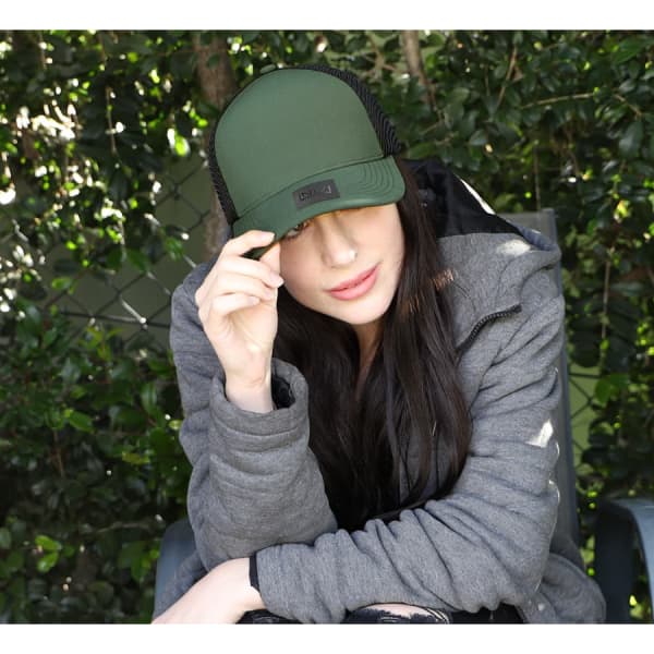 A person wearing a green trucker cap with mesh panels, seated amidst greenery, in a casual grey jacket.