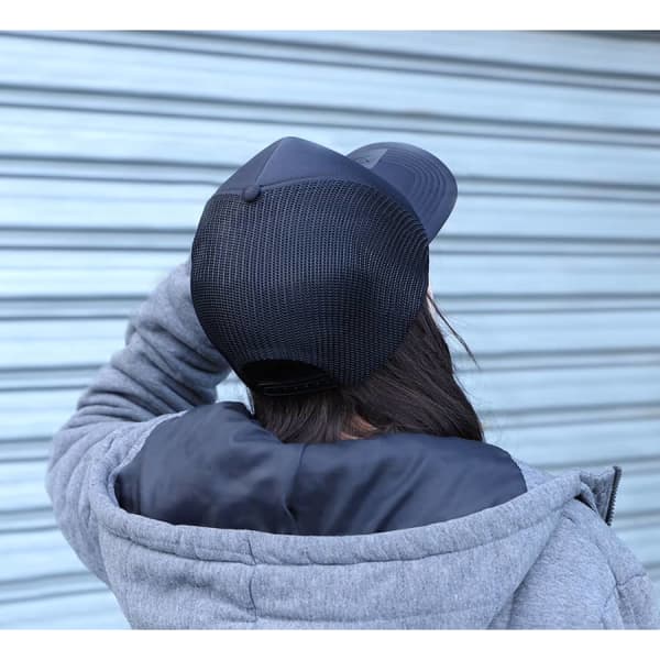 A black trucker cap with a mesh back, worn by a person in a grey hoodie. The background features a blue-grey wall.