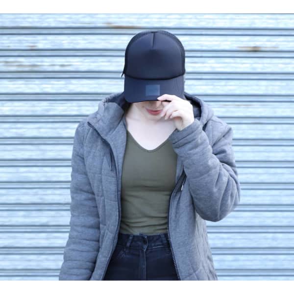 A black trucker cap with mesh detailing, worn by a person in a grey jacket and olive green top, standing against a striped background.