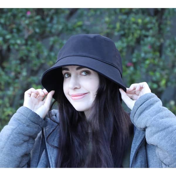 A black bucket hat featured on a model with long dark hair, set against a green backdrop. The hat has a logo on it.