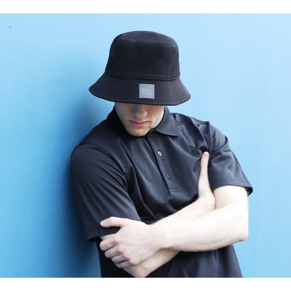 A black bucket hat with a logo, worn by a person in a navy polo shirt against a blue background.