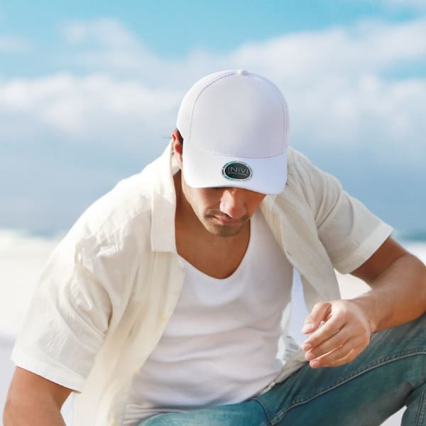 A white cap is worn by a man sitting on the beach, with a logo visible on the front. The scene features a blue sky and ocean.