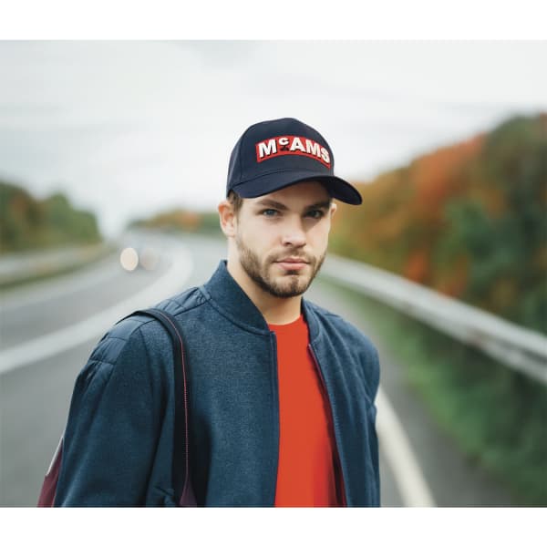 A navy twill cap with a logo, worn by a man in a grey jacket, standing on a road with autumn foliage in the background.